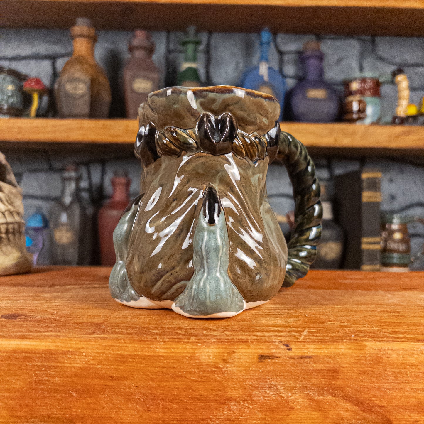 Three-quarter right side view of a handmade Coin Bag Mug held by a dragon hand with two greenish-blue fingers and dark brown nails. The mug has a detailed design featuring a greenish-brown coin bag with a blue-green braided rope handle.