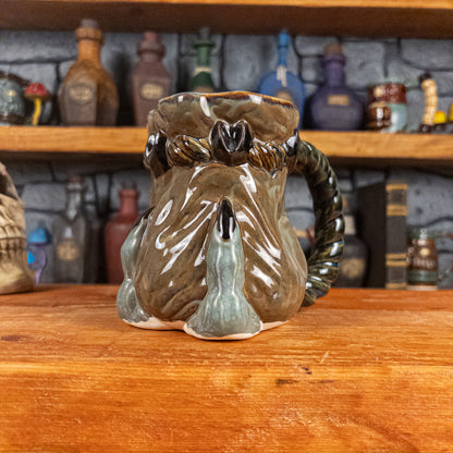 Three-quarter right side view of a handmade Coin Bag Mug held by a dragon hand with two greenish-blue fingers and dark brown nails. The mug has a detailed design featuring a greenish-brown coin bag with a blue-green braided rope handle.