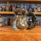 Right side view of a handmade Coin Bag Mug held by a dragon hand with one greenish-blue finger with a dark brown nail. The mug has a detailed design featuring a greenish-brown coin bag with a blue-green braided rope as a handle.