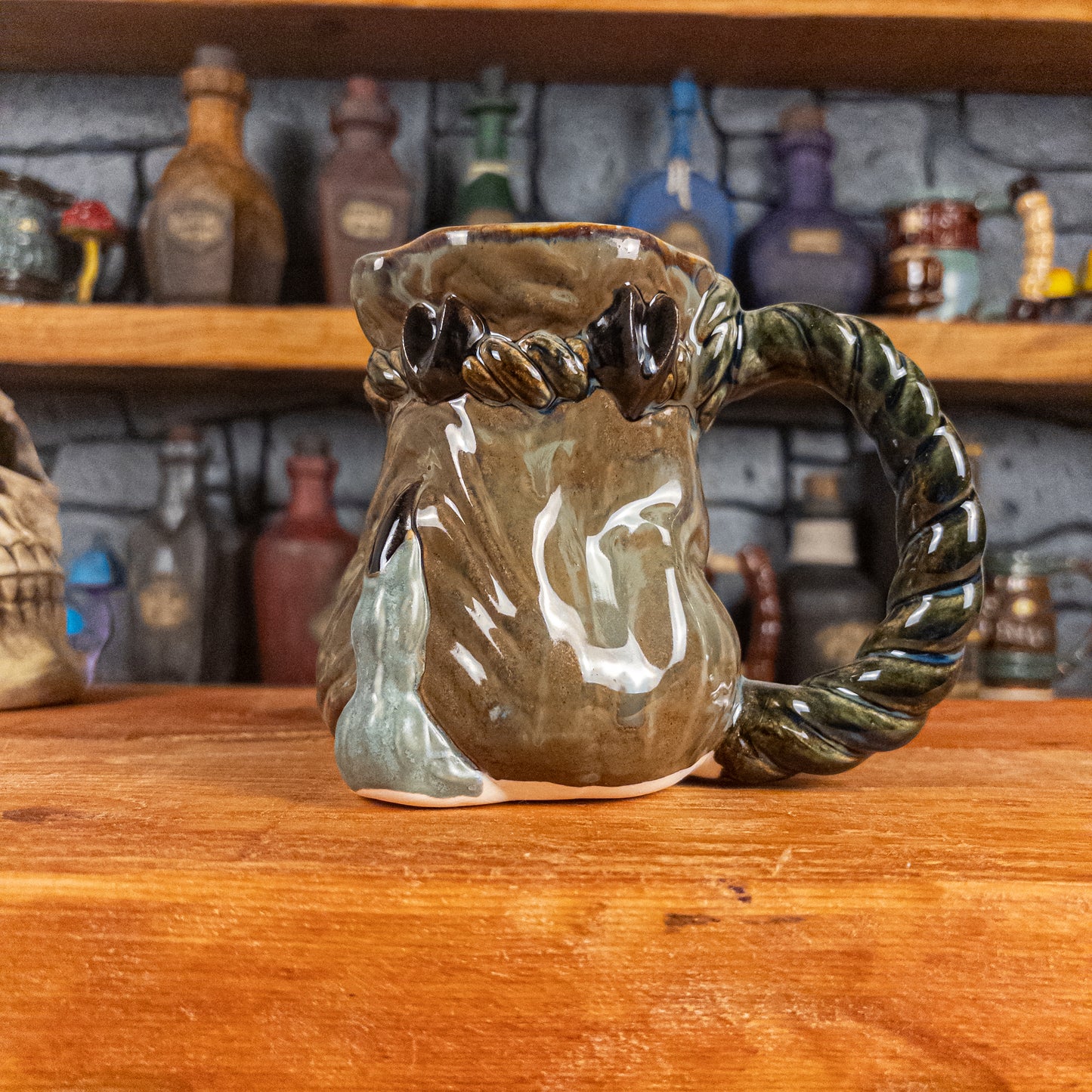 Right side view of a handmade Coin Bag Mug held by a dragon hand with one greenish-blue finger with a dark brown nail. The mug has a detailed design featuring a greenish-brown coin bag with a blue-green braided rope as a handle.