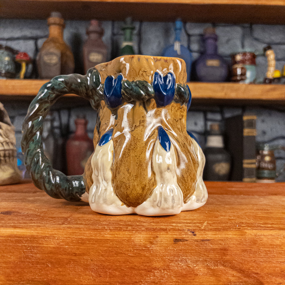 Left side view of a handmade Coin Bag Mug held by a dragon hand with three beige fingers and royal blue nails. The mug has a detailed design featuring a dark gold coin bag with a dark green braided rope as a handle.