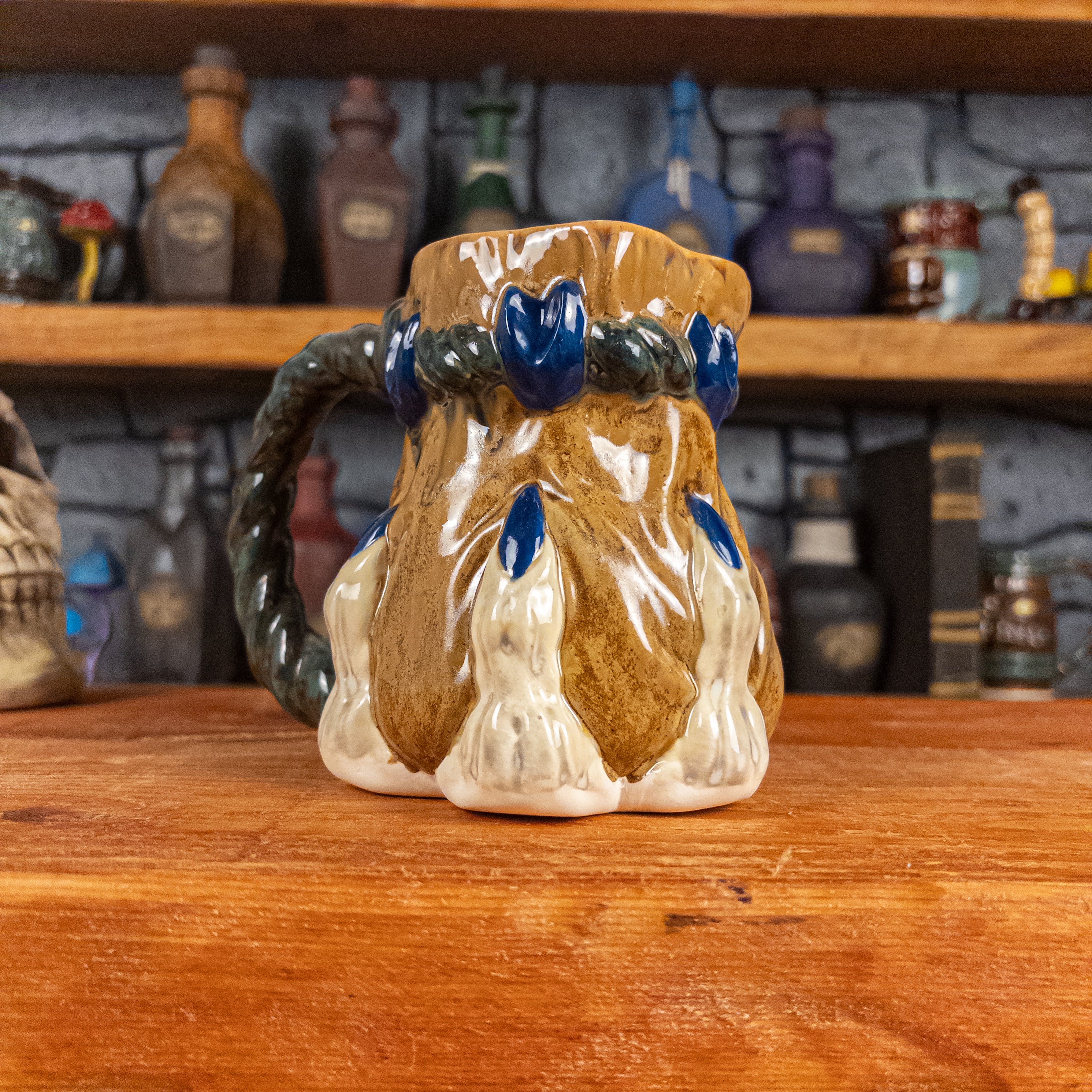 Three-quarter left side view of a handmade Coin Bag Mug held by a dragon hand with three beige fingers and royal blue nails. The mug has a detailed design featuring a dark gold coin bag with a dark green braided rope as a handle.