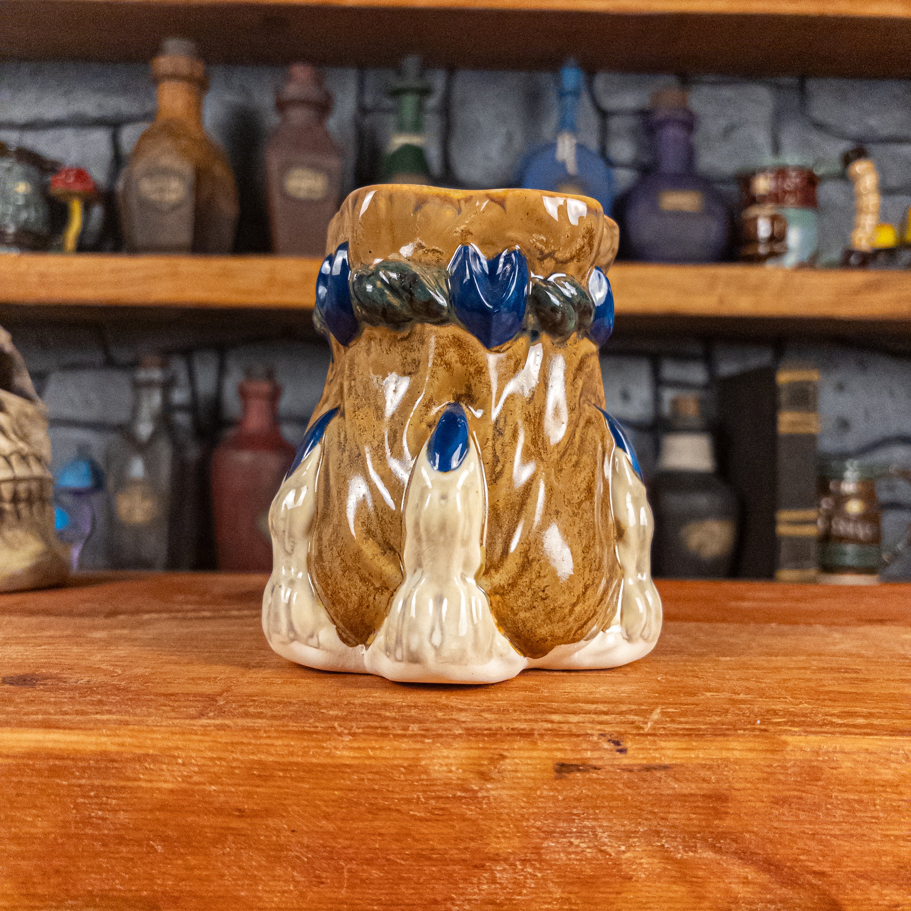 Front view of a handmade Coin Bag Mug held by a dragon hand with three beige fingers and royal blue nails. The mug has a detailed design featuring a dark gold coin bag with a dark green braided rope as a handle.