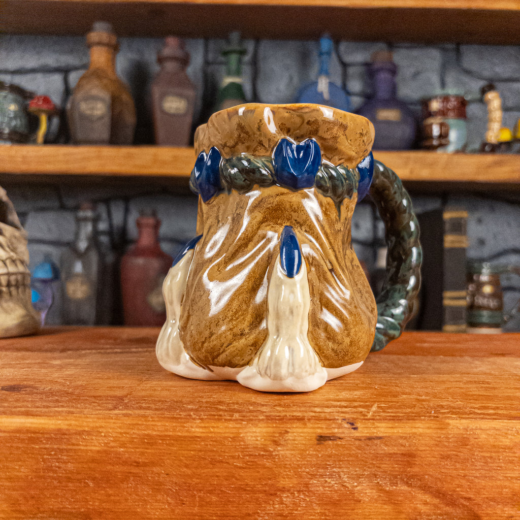 Three-quarter right side view of a handmade Coin Bag Mug held by a dragon hand with two beige fingers and royal blue nails. The mug has a detailed design featuring a dark gold coin bag with a dark green braided rope as a handle.