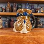 Three-quarter right side view of a handmade Coin Bag Mug held by a dragon hand with two beige fingers and royal blue nails. The mug has a detailed design featuring a dark gold coin bag with a dark green braided rope as a handle.