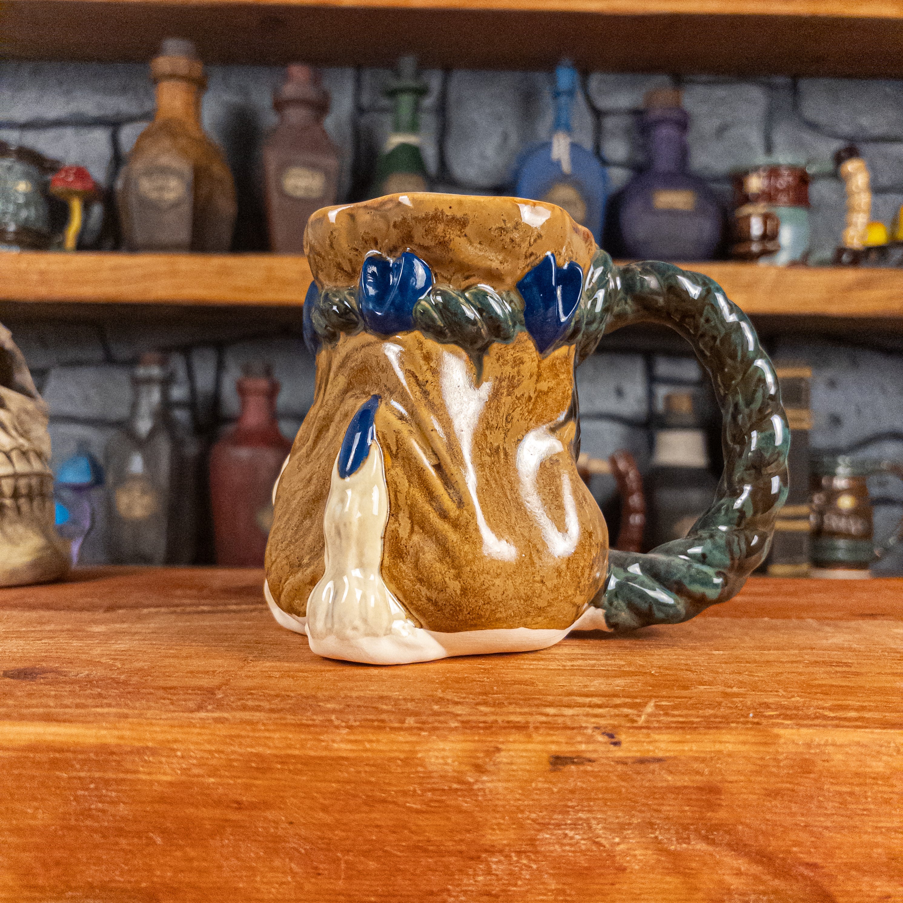 Right side view of a handmade Coin Bag Mug held by a dragon hand with one beige finger and royal blue nail. The mug has a detailed design featuring a dark gold coin bag with a dark green braided rope as a handle.