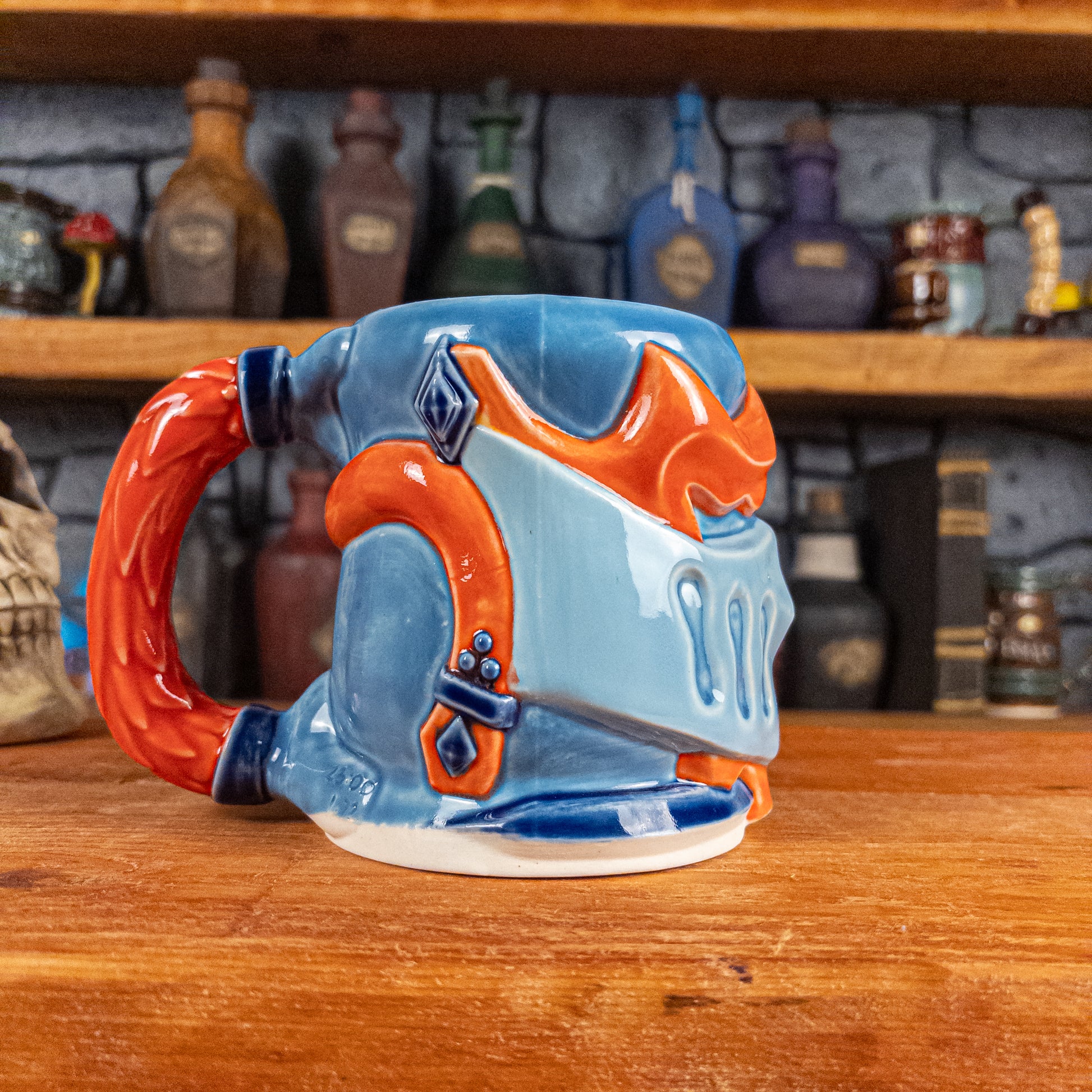 The left side view of a handmade Knight Helmet Mug with a light blue, blue and red-orange armour design, featuring dark blue ornamental rivets and a detailed red handle.
