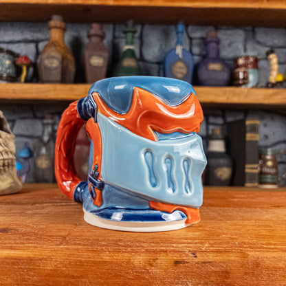 Three-quarter left side view of a handmade Knight Helmet Mug with a light blue, blue and red-orange armour design, featuring dark blue ornamental rivets and a detailed red handle.
