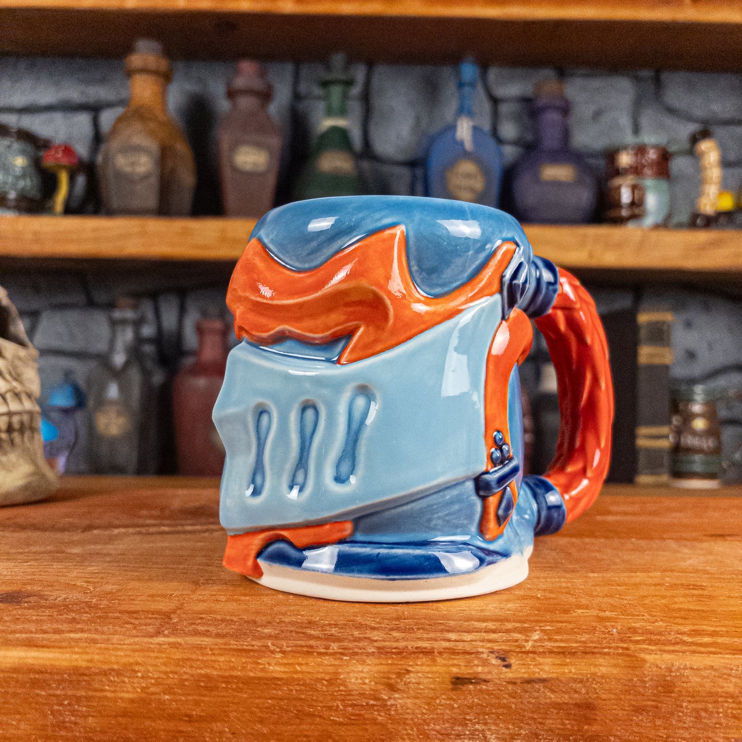 Three-quarter right side view of a handmade Knight Helmet Mug with a light blue, blue and red-orange armour design, featuring dark blue ornamental rivets and a detailed red handle.