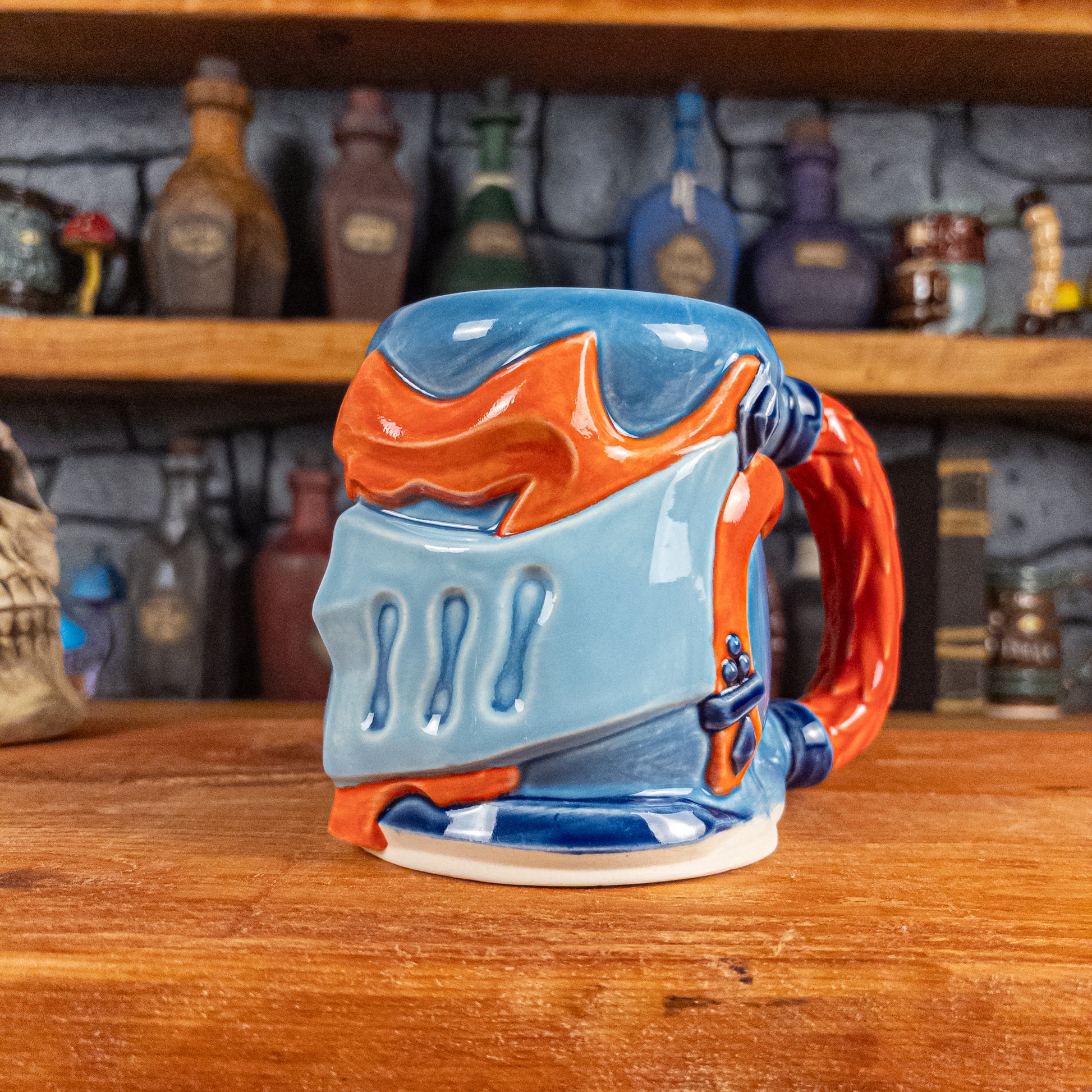 Three-quarter right side view of a handmade Knight Helmet Mug with a light blue, blue and red-orange armour design, featuring dark blue ornamental rivets and a detailed red handle.