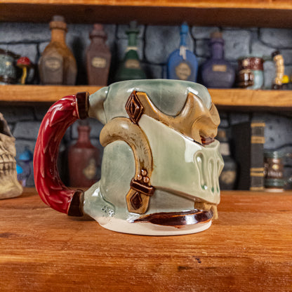 The left side view of a handmade Knight Helmet Mug with a beige and semi-transparent green armour design and brown eye slits, featuring ornamental rivets and a detailed red handle.