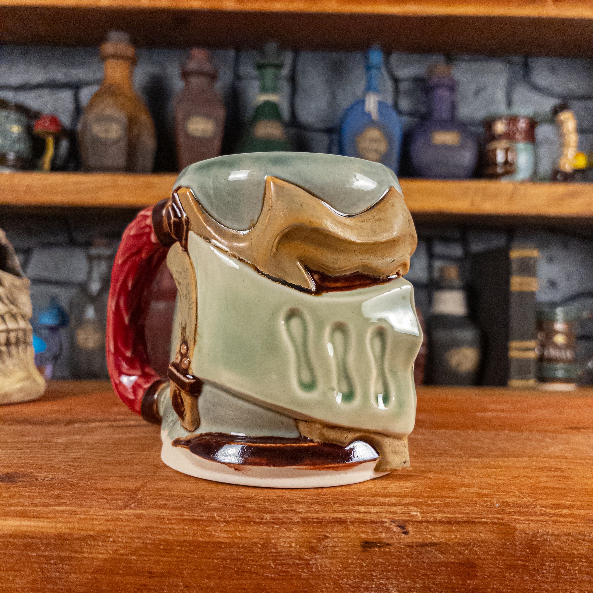 Three-quarter left side view of a handmade Knight Helmet Mug with a beige and semi-transparent green armour design and brown eye slits, featuring ornamental rivets and a detailed red handle.