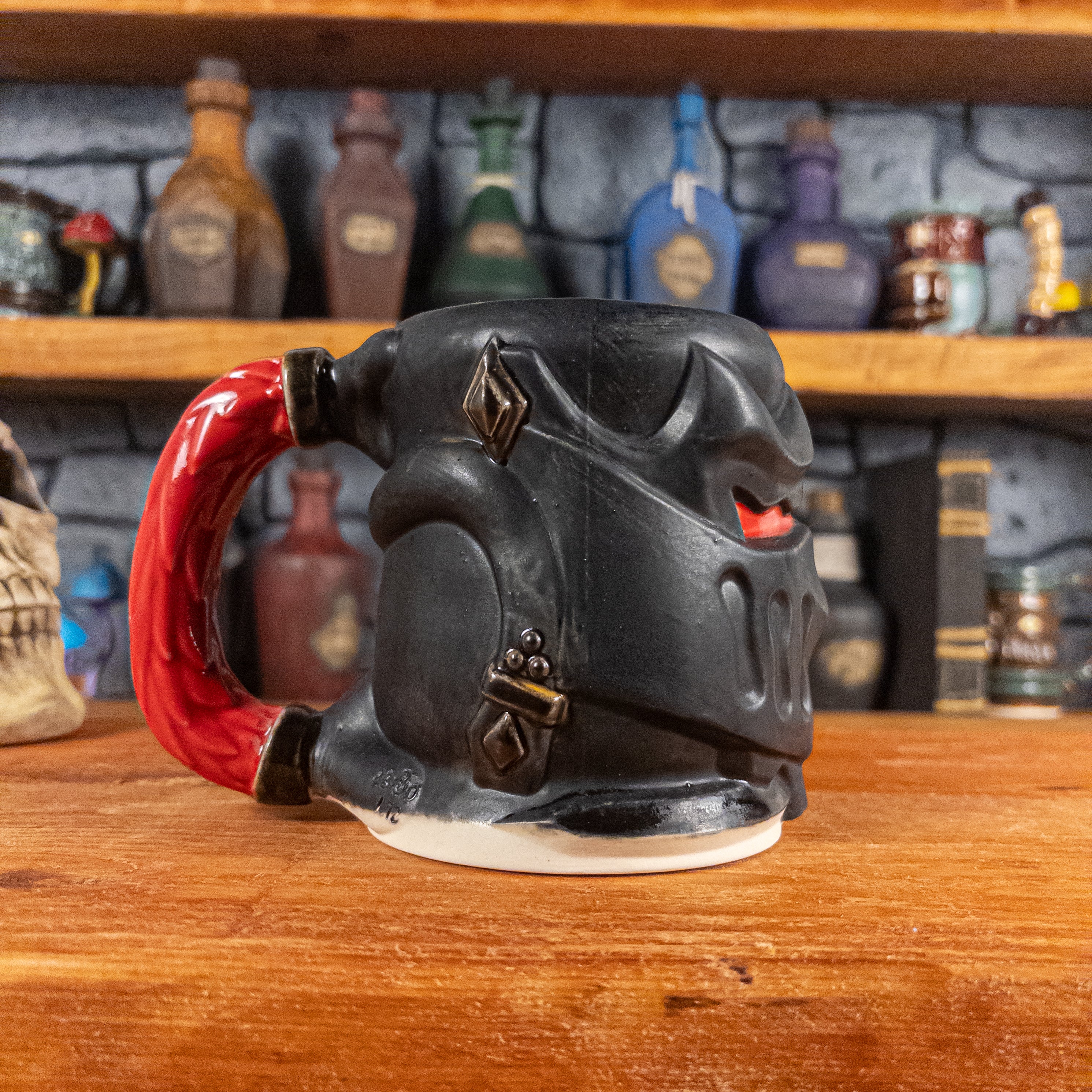 The left side view of a handmade Knight Helmet Mug with a matte black armour design and glossy red eye slits, featuring ornamental rivets and a detailed red handle.