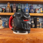 Three-quarter left side view of a handmade Knight Helmet Mug with a matte black armour design and glossy red eye slits, featuring ornamental rivets and a detailed red handle.