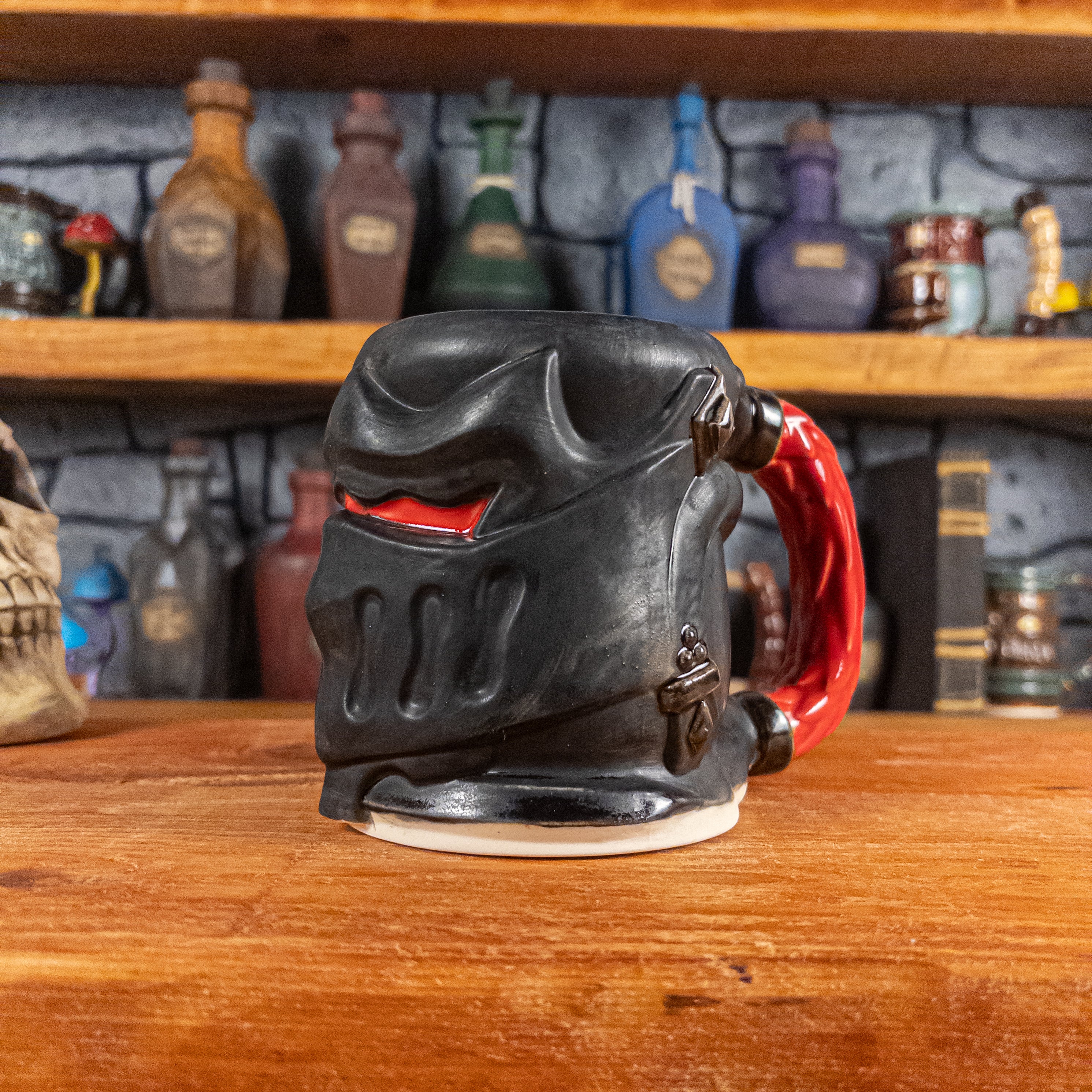 Three-quarter right side view of a handmade Knight Helmet Mug with a matte black armour design and glossy red eye slits, featuring ornamental rivets and a detailed red handle.