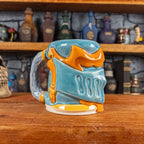 Three-quarter left side view of a handmade Knight Helmet Mug with a light blue and orange armour design, featuring ornamental rivets and a detailed light dusty blue handle.