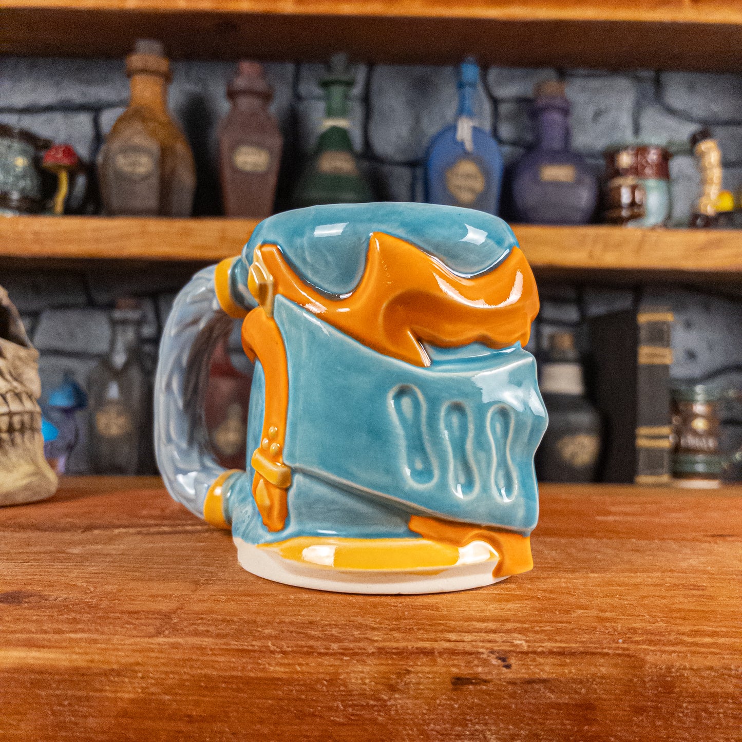 Three-quarter left side view of a handmade Knight Helmet Mug with a light blue and orange armour design, featuring ornamental rivets and a detailed light dusty blue handle.