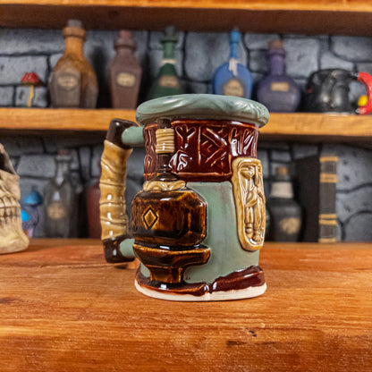 Three-quarter left side view of a handmade Dwarven Mug designed to look like a greenish-blue and dark red barrel with intricate rune carvings, a dark beige dwarf face, and a hammer and anvil design, showing the detailed handle.