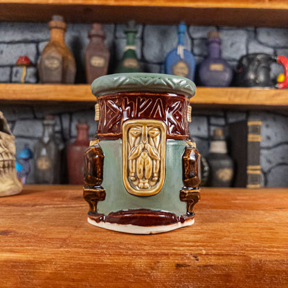 Front view of a handmade Dwarven Mug designed to look like a greenish-blue and dark red barrel with intricate rune carvings, a dark beige dwarf face, and a partial hammer and anvil design.