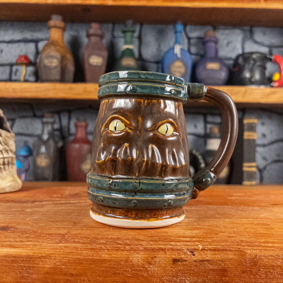 Front view of a handmade Mimic Mug designed to look like a brown wooden barrel with dark brown bands, featuring menacing yellow eyes and teeth. The mug has a handle.