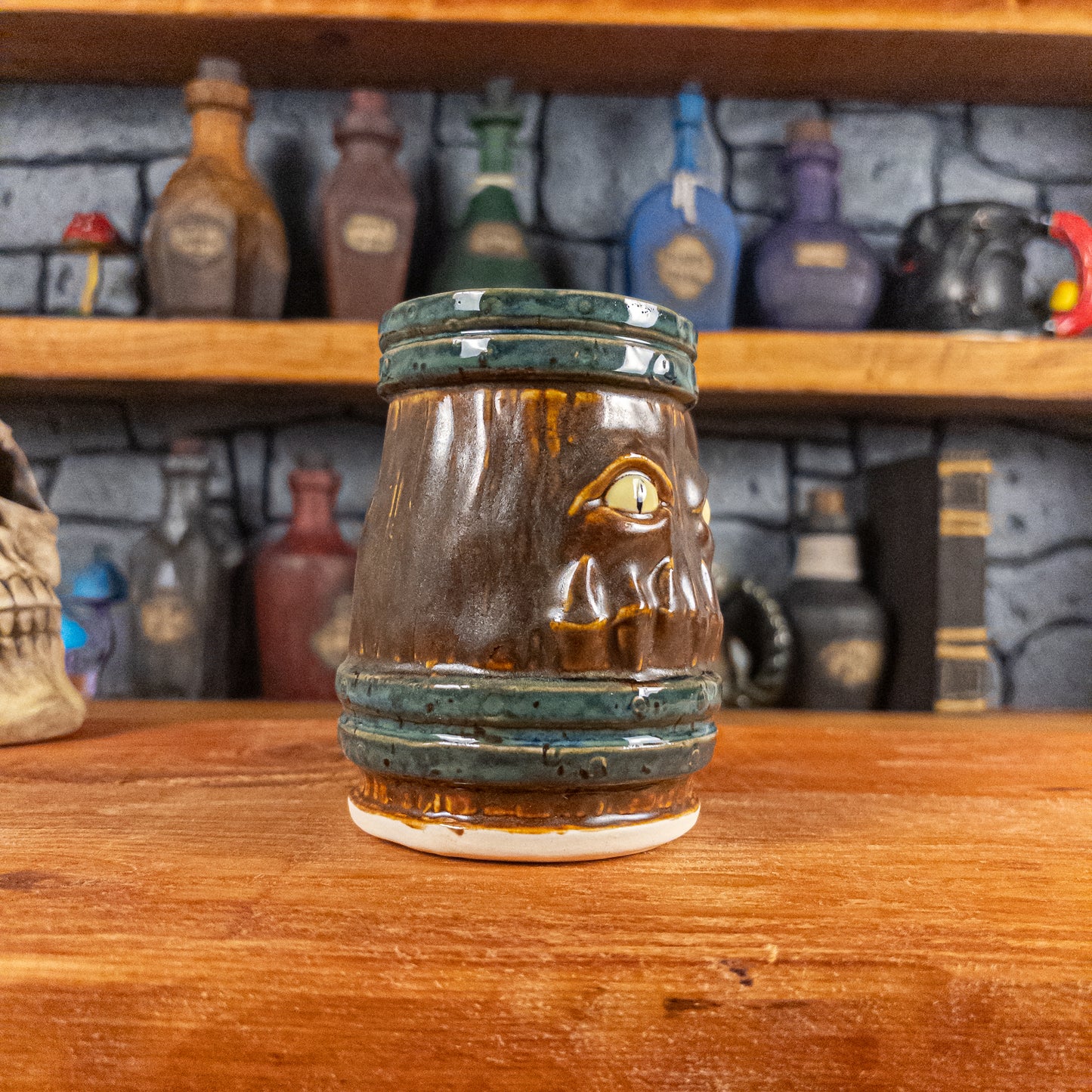 Three-quarter left side view of a handmade Mimic Mug designed to look like a brown wooden barrel with dark green bands, displaying menacing yellow eyes and teeth, and a handle.
