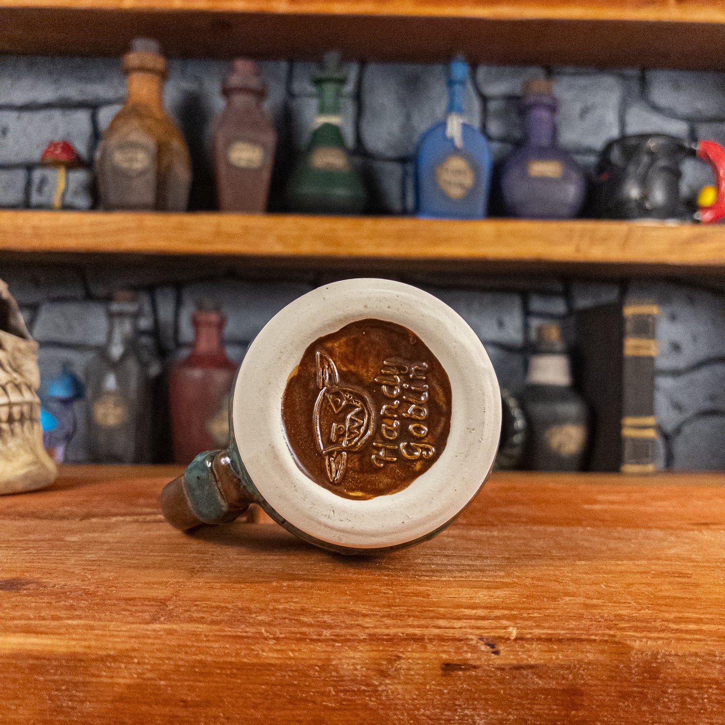 Bottom view of a handmade Mimic Mug with the Handy Goblin logo embossed in the center, featuring a speckled brown base.