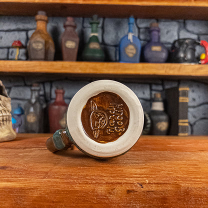 Bottom view of a handmade Mimic Mug with the Handy Goblin logo embossed in the center, featuring a speckled brown base.