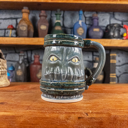 Front view of a handmade Mimic Mug designed to look like a greenish-blue wooden barrel with dark brown bands, featuring menacing yellow eyes and teeth. The mug has a handle.