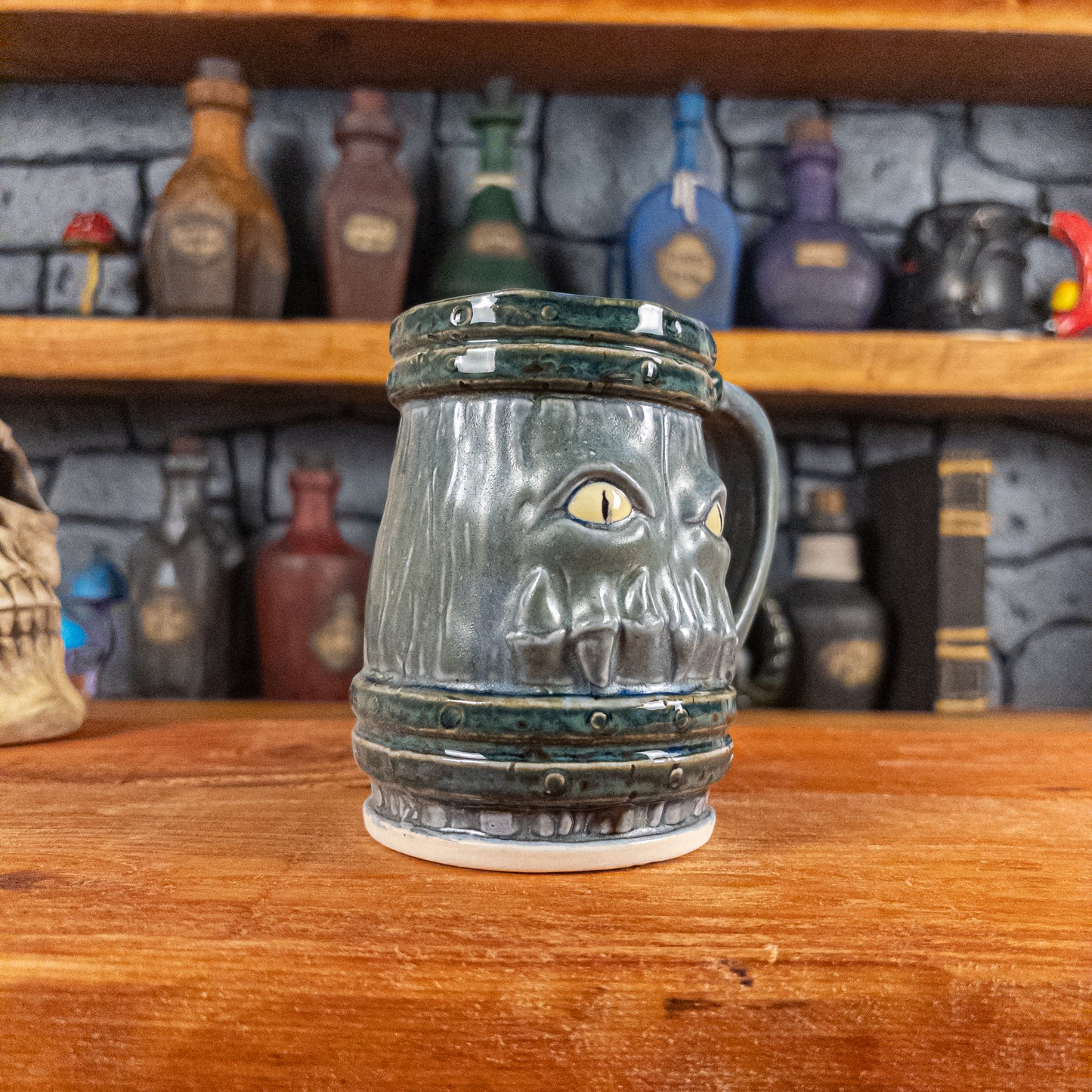 Three-quarter left side view of a handmade Mimic Mug designed to look like a greenish-blue wooden barrel with dark green bands, showing menacing yellow eyes and teeth, and a handle.