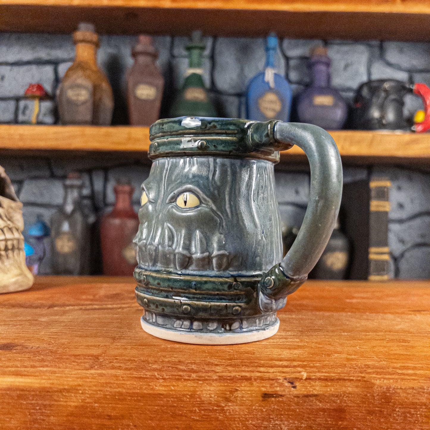 Three-quarter right side view of a handmade Mimic Mug designed to look like a greenish-blue wooden barrel with dark green bands, showing menacing yellow eyes and teeth, and a handle.