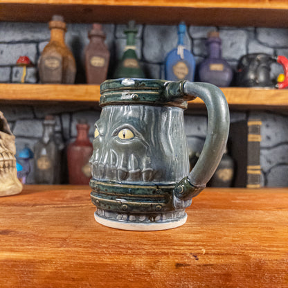 Three-quarter right side view of a handmade Mimic Mug designed to look like a greenish-blue wooden barrel with dark green bands, showing menacing yellow eyes and teeth, and a handle.