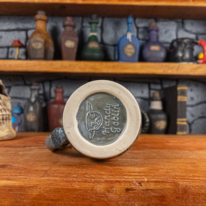 Bottom view of a handmade Mimic Mug with the Handy Goblin logo embossed in the center, featuring a greenish-blue base.