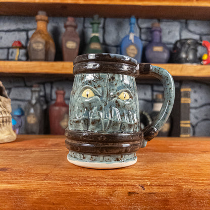 Front view of a handmade Mimic Mug designed to look like a speckled light blue wooden barrel with dark brown bands, featuring menacing yellow eyes and teeth. The mug has a handle.