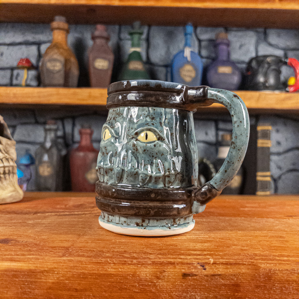 Three-quarter right side view of a handmade Mimic Mug designed to look like a speckled light blue wooden barrel with dark brown bands, displaying menacing yellow eyes and teeth, and a handle.