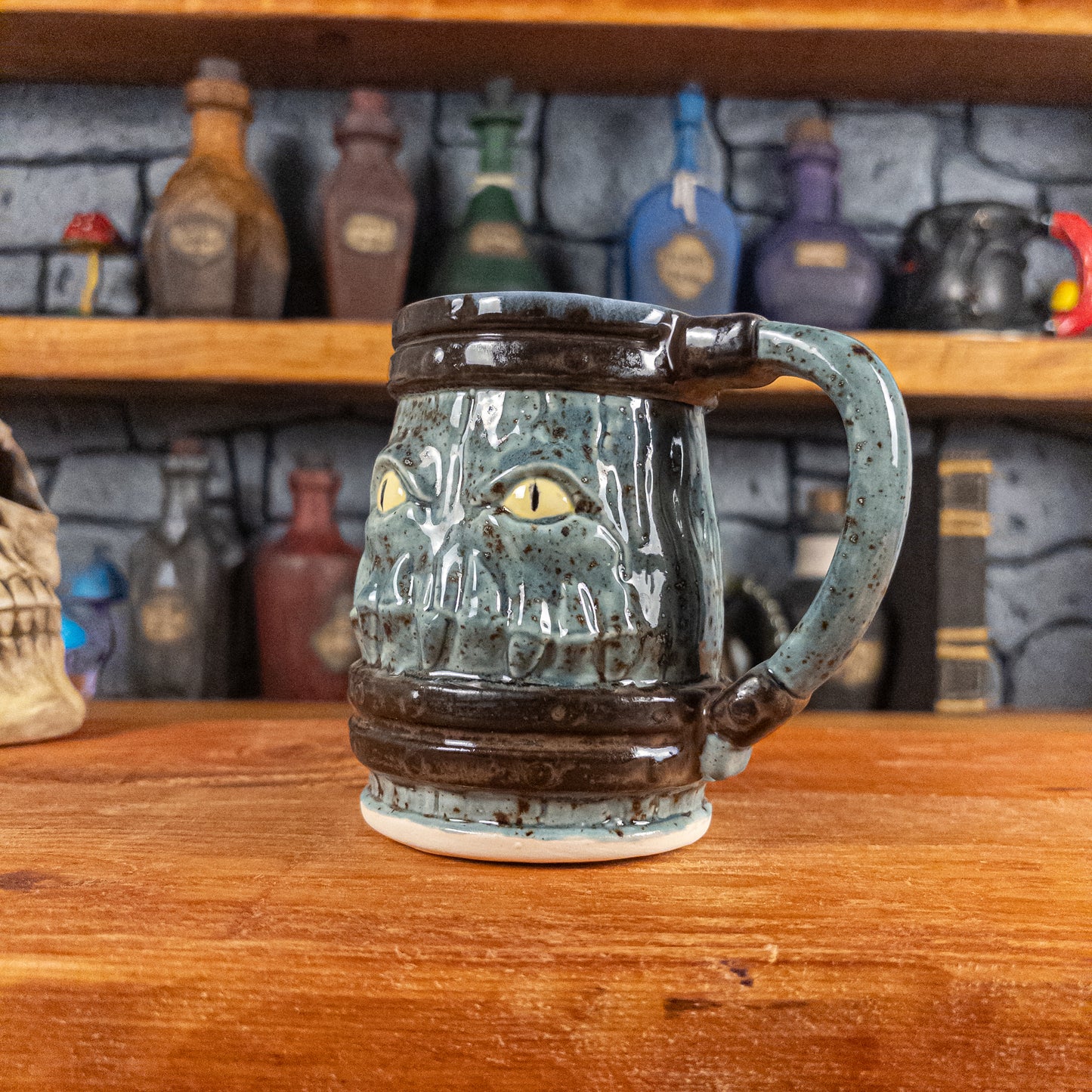 Three-quarter right side view of a handmade Mimic Mug designed to look like a speckled light blue wooden barrel with dark brown bands, displaying menacing yellow eyes and teeth, and a handle.
