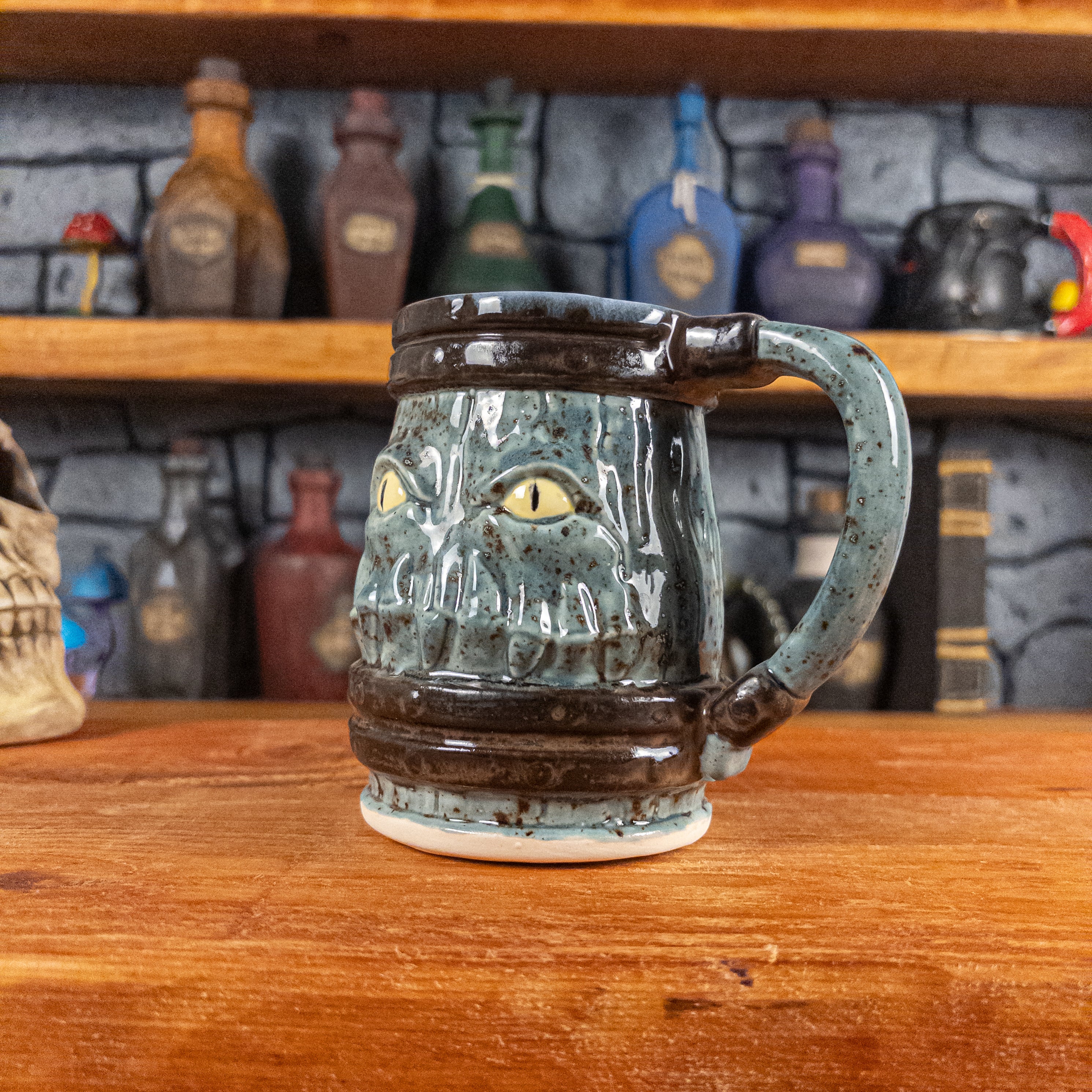 Three-quarter right side view of a handmade Mimic Mug designed to look like a speckled light blue wooden barrel with dark brown bands, displaying menacing yellow eyes and teeth, and a handle.