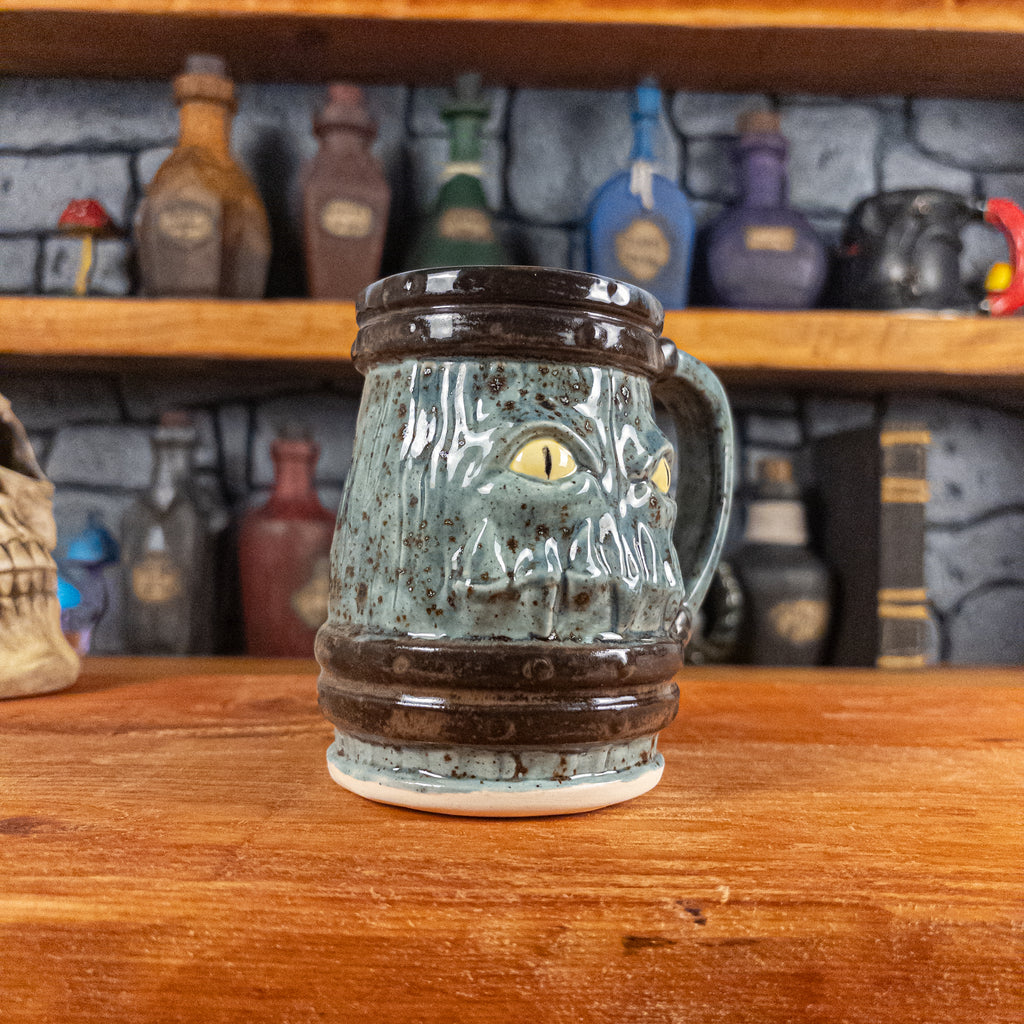 Three-quarter left side view of a handmade Mimic Mug designed to look like a speckled light blue wooden barrel with dark brown bands, showing menacing yellow eyes and teeth, and a handle.