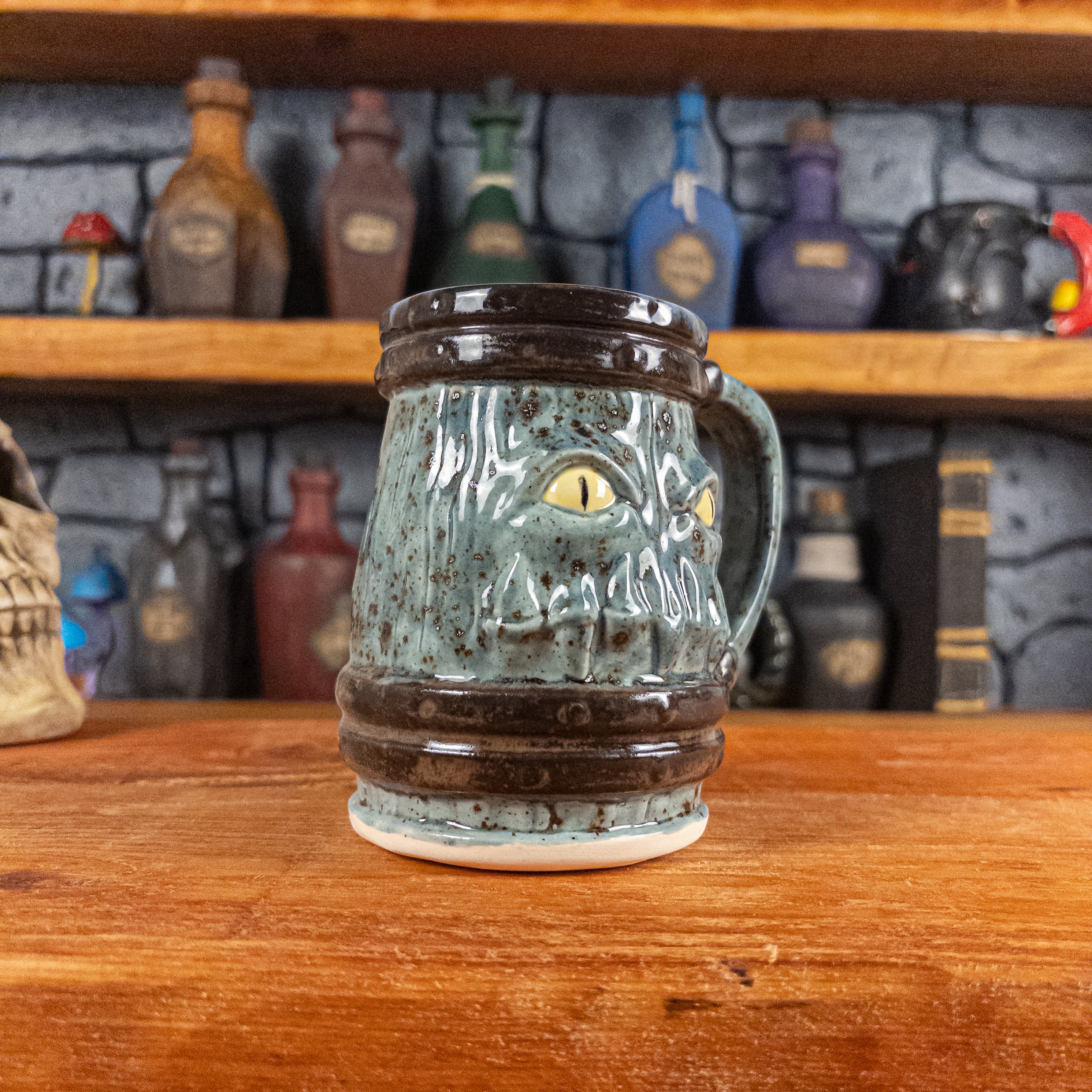 Three-quarter left side view of a handmade Mimic Mug designed to look like a speckled light blue wooden barrel with dark brown bands, showing menacing yellow eyes and teeth, and a handle.