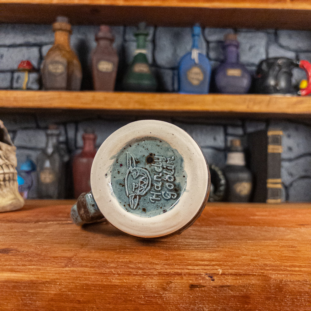 Bottom view of a handmade Mimic Mug with the Handy Goblin logo embossed in the center, featuring a speckled light blue base.