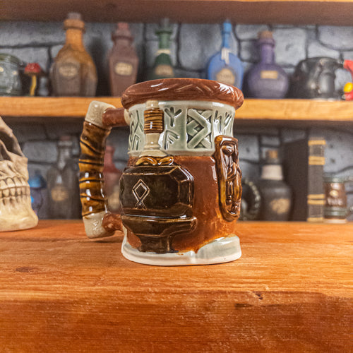 Three-quarter left side view of a handmade Dwarven Mug designed to look like a light green and reddish-brown barrel with intricate rune carvings, a dwarf face, and a hammer and anvil design, showing the detailed handle.